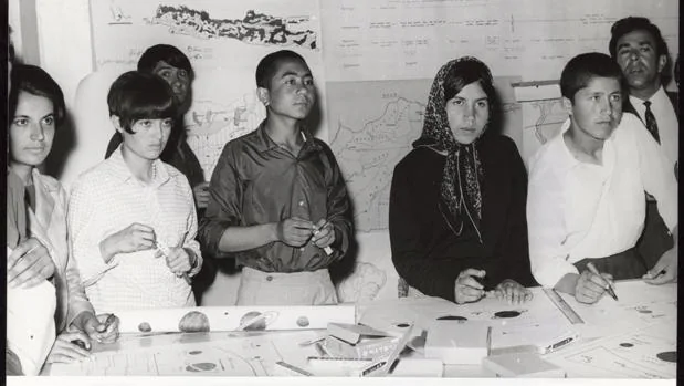 Un grupo de estudiantes (tanto mujeres como hombres) están de pie junto a una mesa en un aula escuchando una clase en Kabul, Afganistán, en 1968